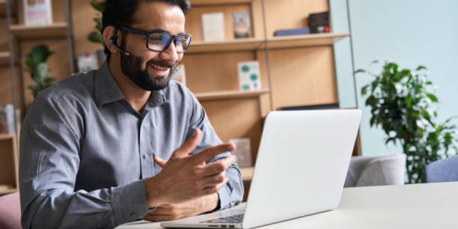 Happy indian business man remote teacher, customer support manager wearing headset talking at virtual meeting consulting client on video call giving distance learning class at home office call center.